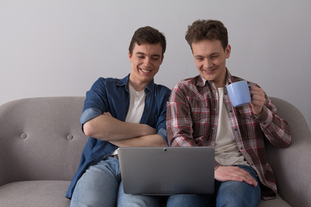 Couple of teens looking down at computer screen sitting on couch. Two friends wearing jeans and button up shirts relaxing at home on sofa watching film on computer.の写真素材