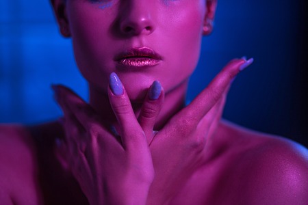 The model in neon tones with pronounced lips holds hands on the face. Sexy girl model with bare shoulders. Close Up Shoot.の写真素材