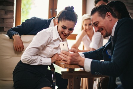 Handsome Businessman Shows Female Colleague Some Information On His Phone. Smiling Woman In Formal Cloth Looks On The Smartphone Of Her College. Teamwork Business Meeting.の写真素材
