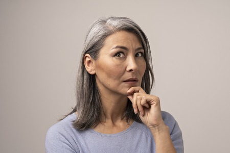 Mongolian-Looking Woman with Gray Hair on a Gray Background. Her Face Looks Thoughtful. The Woman s Gaze is Directed Upwards. Her Hand Props Her Chin. Close Up Shoot.の写真素材