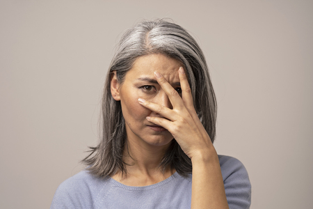 Sad Mongolian Woman on Gray Background. She Has Gray Hair. She Covers Her Sad Face with Her Hand. Close Up Shoot.の写真素材