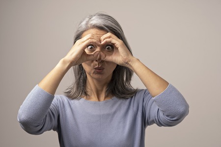 Mongolian Mature Woman Holds Hands As if Looking Through Binoculars. The Woman Has Gray Hair. Her Face Looks Funny. She Opened Her Eyes Wide with Surprise. Behind the Woman is a Gray Background. Close Up Shoot.の写真素材