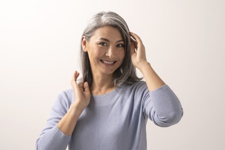 Charming Woman Gently Touches Her Grey Hair. Elderly Smiling Asian Woman Posing In The Studio....