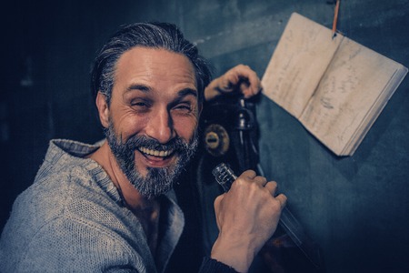 Handsome Man Laughs At Camera While Holding A Bottle Of Whiskey. Drunken Bearded Man s Hand Rests On The Rotary Phone As He Laughs Out Loud And Drinks. Blurred Image Of A Notebook Hanging On The Wall Near The Rotary Phone. Portrait.の写真素材