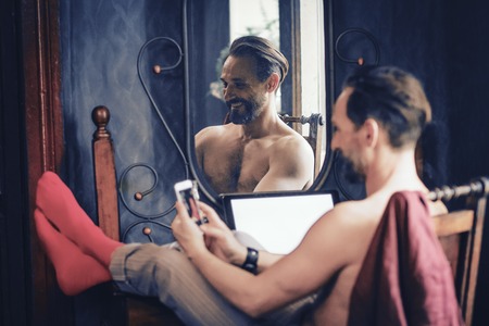 A Handsome Man With a Beard is Sitting at His Desk at Home. Throwing His Legs on the Table and Leafing Through the Phone He Laughs. A Man With a Naked Torso Looks Brutal. Close Up Shot.の写真素材