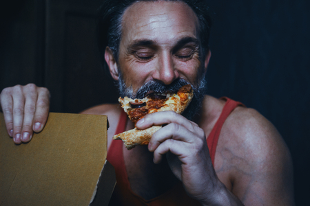 Handsome Bearded Man Bites A Big Piece Of Pizza. Happy Face Of Satistified Man Biting A Piece Of Pizza. Hungry Man Eats Delivered Pizza. Portrait. Closeup. Eating Pizza Conceptの写真素材