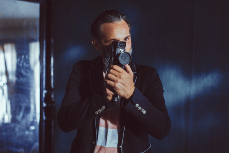 A Man Moviemaker Shoots With a Vintage Camera. A Beard and a Black Jacket on it Makes it Solid. The Man is in a Dark Room.の写真素材