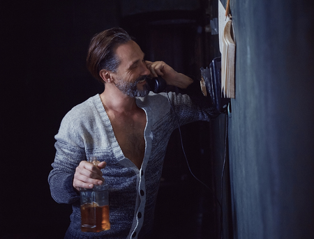 A 40 Year Old Man Loves to Drink Alcohol. He Looks Brutal With a Beard and a Bottle in His Hand. He Calls On An Old Telephone and is Joking at Someone.の写真素材