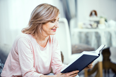 Lovely Woman Reads A Book Sitting On The Sofa. Pretty Mature Lady Is Fully Immersed Into The Book. Side View. Blurred Backgroundの写真素材