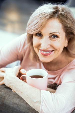 Smiling Woman Looks Over 50 Years Old. She is Resting in Her Living Room on the Couch. In the Hands of a Woman a Pink Cup of Tea. Close Up Shot.の写真素材