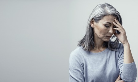 Serious Middle-Aged Woman Touches Her Forehead and Thinking About Something. Upseted Asian Woman With Grey Hair Stands And Touches Her Forehead Thinking. Right Side Portraitの写真素材