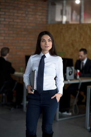 Attractive brunette is an office worker who is standing in office, looking on the right, holding a laptop, she is wearing a white shirt and a black trousers. Two colleguas are working behind her.の写真素材