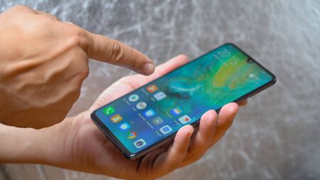 Man holding Huawei Mate 20 X smartphone in one hand and pointing with finger on screen on shining grey background. Display is turned on and main menu is seen Kyiv, Ukraine 15th of June 2019のeditorial素材