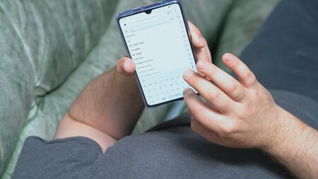 Man lying on couch and holding Huawei Mate 20 X smartphone in one hand. He is typing some text in...