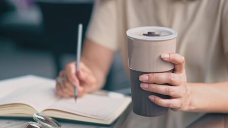 The Middle-Aged Asian Woman Is Sitting At The Table And Tapping Her Notebook With A Pencil.The Business Lady Is Also Holding A Carton Coffee Cup In Her Handの写真素材