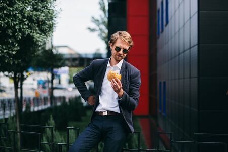 A Male Office Worker In Dark Sunglasses Sits Outside In The Summer And Eats A Burger At Lunchtime. A Young Man Rushing To Eat His Food And Hurries To Work. Business Concept Photoの写真素材