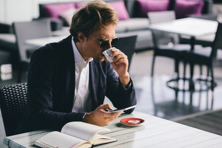 An Office Worker In Sunglasses And A Blue Formal Suit Went To A Restaurant To Drink Some Coffee And Write Something In His Notebook. Attractive Freelancer Guy Working Remotely. Business Concept Photoの写真素材