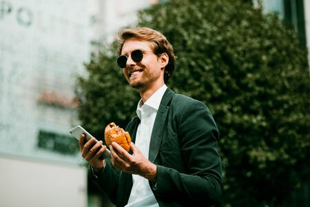 Good-looking Businessman Man In A Dark Suit And Black Sunglasses Is Sitting On the Metal Fence In The City Street And Having His Lunch. He Is Holding A Smartphone In His Hand. Toned image.の写真素材