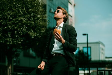 Nice-looking Businessman Man In A Dark Suit And Black Sunglasses Is Sitting On the Metal Fence In The City Street And Having His Lunch. He Is Looking Up. Toned image.の写真素材
