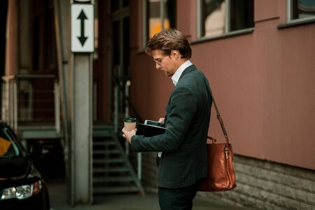 Well-dressed Young Man Is Looking At The Screen Of His Phone And Holding Notebook And A Carton...