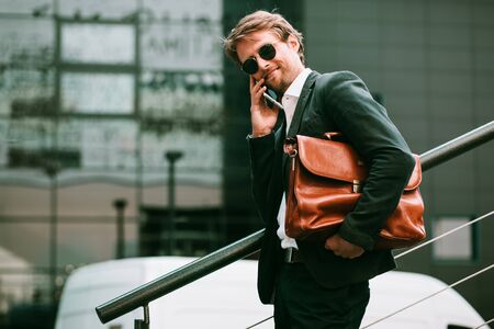 The Smartly-dressed Man Is Standing On The Stairs In The City Center. He Is Holding A Brown Briefcase Under His Arm and Speaking On His Phone. Toned image.の写真素材