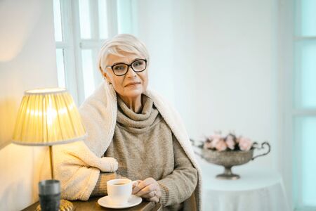 Old Woman In A Woolen Sweater And A Plaid On Her Shoulders Is Standing Near The Cupboard.の写真素材