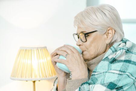 Granny Is Drinking Tea. Senior Woman Wearing Glasses Is Wrapped In A Checkered Plaid To Feel Comfortable. Switched-on Lamp Is Next To Her. Back Side Shotの写真素材