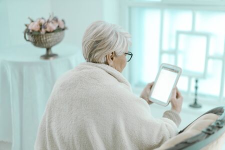 Old Woman Wrapped In A Warm Plaid Sits In Her Cosy Arm-chair Near The Big Window. She Is Reading Something Intresting On Her Tablet. Vase With Flowers Is Standing By Her. Back Side Shot.の写真素材