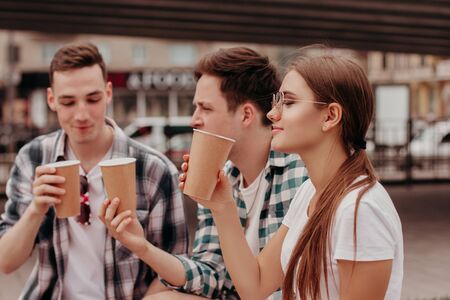Three Friends Having Hot Drinks outdoors In Plastic Cups Outdoor. The Girl Is Smelling The Drink And...