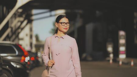 A businesswoman confidently walks through supermarket car park on sunny day. An Asian brunet woman goes through the parking lot and happily smiling.の写真素材