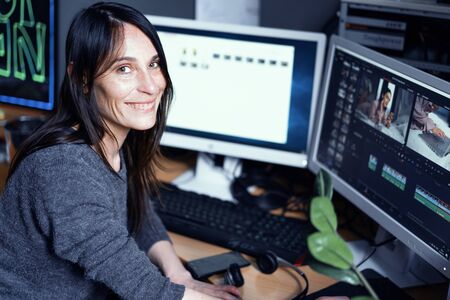 A Young Dark-haired Female Video Maker Smiles And Works At Computer In A Photo Studio. Freelancer Girl Is Doing Some Video Processing. Computer Work Concept Photoの写真素材