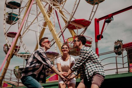 Young People Have Fun On Vacation In The Park. Two Beautiful Guys And A Girl Rest And Hold Hands...