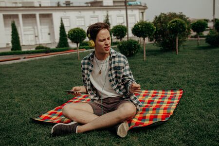 Teenager Enjoys Music With Headphones Sitting On Multi-Colored Mat In A City Park In Summer Day.