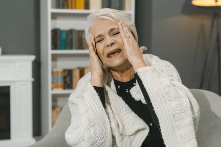 Gray-Haired Elderly Lady Touches Her Temples With Her Hands While Sitting In An Armchair. She Closed Her Eyes And Opened Her Mouth With A Sigh. Cozy Home Interior On A Blurred Background. Toned Imageの写真素材