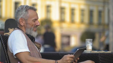 Intelligent bearded man holds a tablet and laughs in a cozy street cafe. Longboard and a drinking cup of coffee. Urban architecture in the backgroundの写真素材