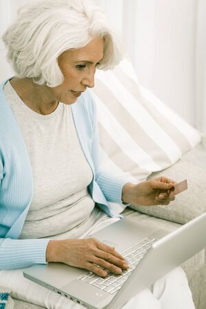 gray haired senior italian woman holds credit or debit card and use laptop to do online payments, or...