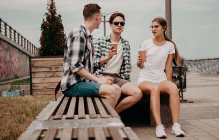 A Group Of Young Students Communicate And Drink From Paper Glasses Sitting On A Bench. Two Guys And A Girl Arranged A Halt While Traveling Through The European City. A Group Of Friends In Kejual And Sneakers Rest And Drink Tea And Coffee.の写真素材