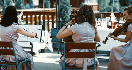 String Orchestra Performing Classical Music Outside Summer Terrace Three Wemen Wearin Lilliac Dresses And Holding Violins Shot From Behind.の写真素材