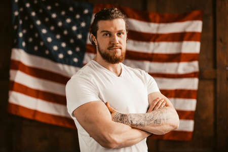Patriotic American With Tattoo On His Hand Standing On The American Flag Background. The Successful Attractive Man Sported Physique In A White T-Shirt Looking Confidentの写真素材