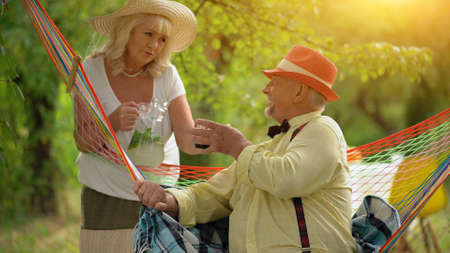 The Elderly Man Is Sitting In The Colourful Hammock Covered With A Blue Blanket.His Wife Is Giving Him A Glass With A Cold Lemonade.They Rest In a Green Gardenの写真素材