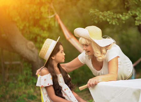 Grandmother speak to little girl sitting on chair in garden. Old lady has conversation with granddaughter outdoor across park.の写真素材