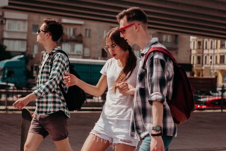Friends Students Walk Around The City During The Summer Holidays. Guys In Plaid Shirts And A Girl In White Walk Along A Busy Street Of A European City. The Girl Dances On The Run.の写真素材