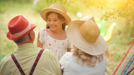 The Grandparents Are Sitting With Their Back In The Hammock.They Are Looking At Their Beautiful Granddaughter.The Girl Is Standing Near Them And Looking At Grandpaの写真素材