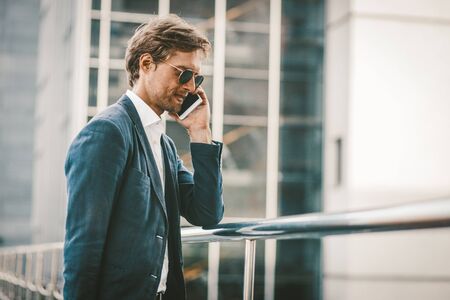 Handsome man walking on a bridge and talking on the phone. Happy smiling young businessman standing outside and having conversation on the phone with urban background.の写真素材