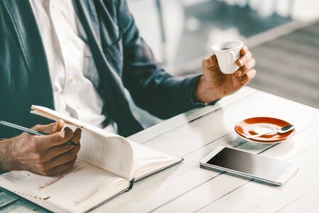 Male businessman working during lunch. Yooung man in business suit with espresso cup in a had doing entries in his schedule book. Man working at restaurant or cafeの写真素材