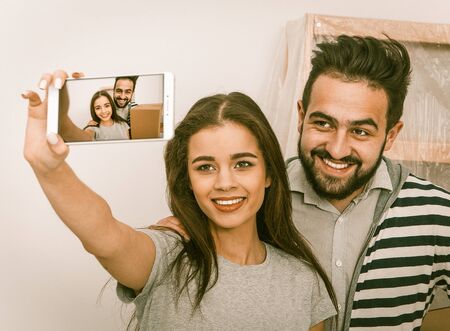 Relocation young happy couple making selfie shot in their new apartment. Man holding boxes smiling...