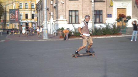 Intelligent light-skinned hipster skates outdoors and looks forward. European man with a beard rushes forward on a longboard on the central city street.の写真素材