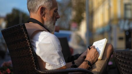 A grey light-skinned bearded man inspiredly writes down ideas in a notebook in a street cafe. Close Up.の写真素材