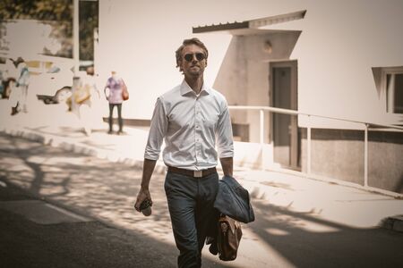 Young handsome businessman going to work, carries a briefcase and jacket in hand and coffee or tea in disposable cup in other hand on urban backgroundの写真素材