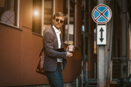 Handsom man in a sute and briefcase looking a side while he crossing a rode. Young businessman holding a disposable cup of coffee and planner.の写真素材